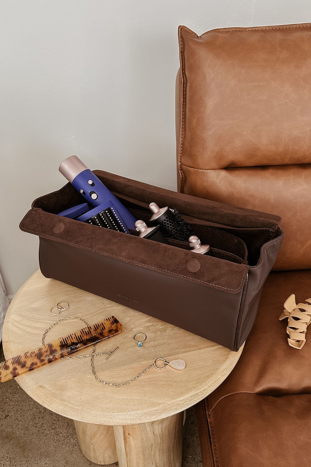 Brown pleather storage case with hair care products on a wooden stool next to a brown leather chair.