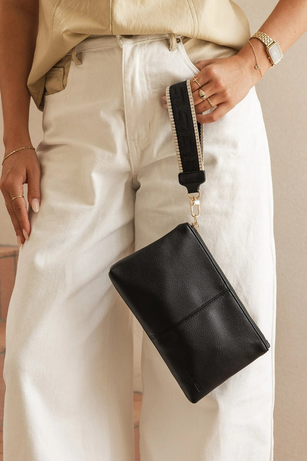 A person wearing cream wide-leg pants and a beige top holds a black leather wristlet clutch with a black strap and gold-tone hardware.