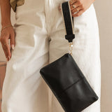 A person wearing cream wide-leg pants and a beige top holds a black leather wristlet clutch with a black strap and gold-tone hardware.