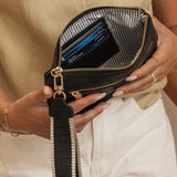 A close-up of a person holding an open black wristlet pouch with a striped black-and-white strap and gold-tone hardware. The pouch contains cards, and the person is wearing a beige top and white pants.