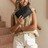 A woman smiling while holding a black leather clutch bag, wearing a beige sleeveless top and white pants, standing in front of a staircase with a wooden handrail.