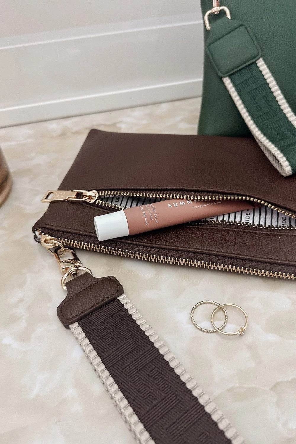A brown leather zip pouch with a matching brown and white patterned strap, shown on a light marble surface next to a pink lip product, two silver rings, and part of a green bag.