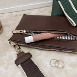 A brown leather zip pouch with a matching brown and white patterned strap, shown on a light marble surface next to a pink lip product, two silver rings, and part of a green bag.