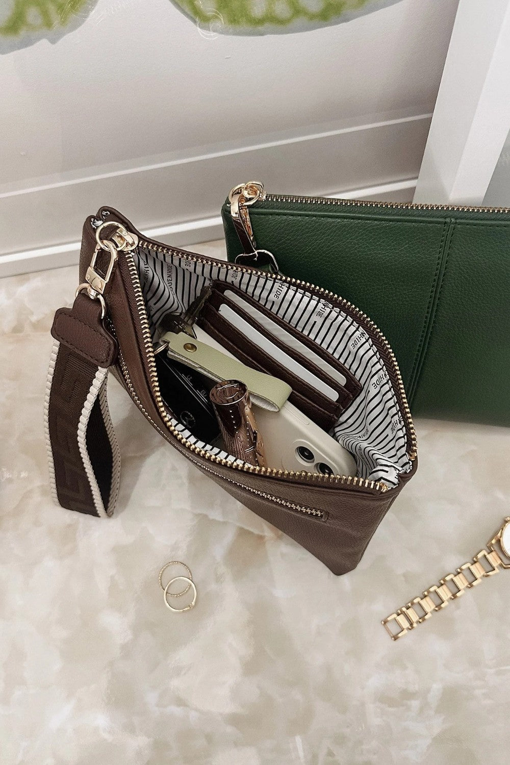 A brown leather zippered clutch wallet shown open, revealing a striped interior lining and compartments filled with cards and a phone. A green wallet and a gold chain bracelet are partially visible in the background on a light marble surface.