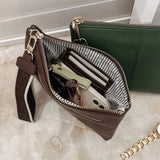 A brown leather zippered clutch wallet shown open, revealing a striped interior lining and compartments filled with cards and a phone. A green wallet and a gold chain bracelet are partially visible in the background on a light marble surface.