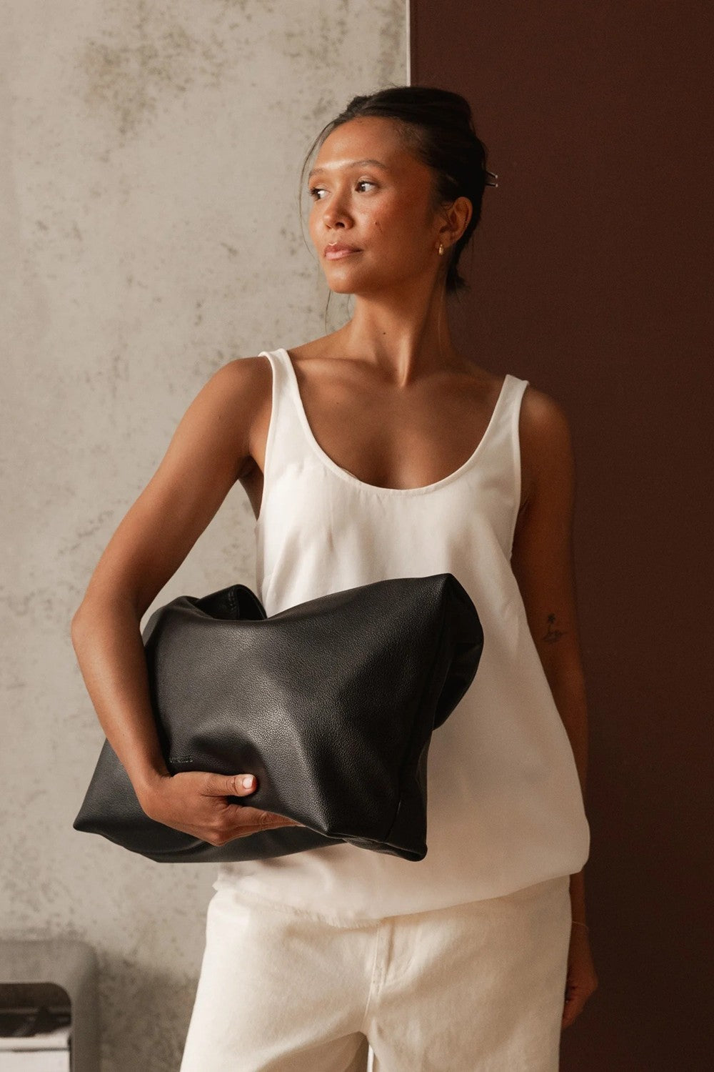 A woman wearing a white tank top and white pants holds a large black leather bag against a split textured gray and dark brown background.