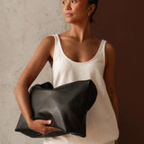 A woman wearing a white tank top and white pants holds a large black leather bag against a split textured gray and dark brown background.