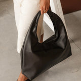 A person wearing white pants and dark flip-flop sandals holds a large black slouchy shoulder bag with a cutout handle in a minimalist indoor setting.