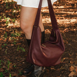 Person holding a burgundy handbag outdoors on a leafy ground