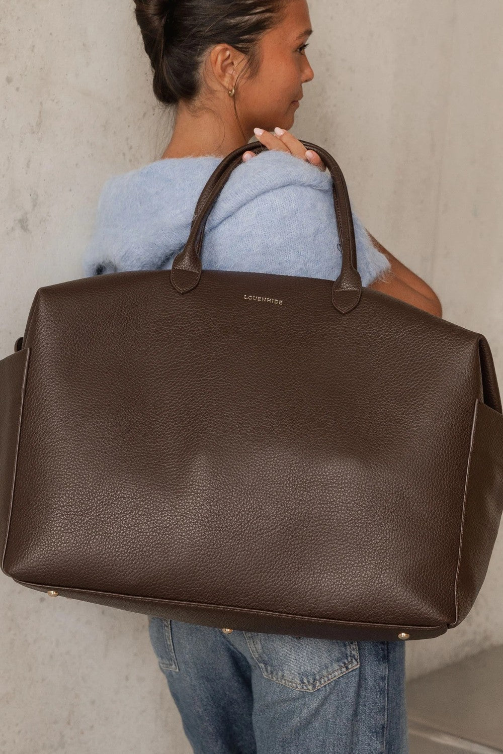 A woman in a light blue sweater and jeans carries a large brown pebbled leather duffel bag with top handles. The brand name 'LOUENHIDE' is printed in gold on the front of the bag, set against a neutral concrete wall background.