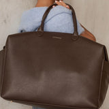 A woman in a light blue sweater and jeans carries a large brown pebbled leather duffel bag with top handles. The brand name 'LOUENHIDE' is printed in gold on the front of the bag, set against a neutral concrete wall background.