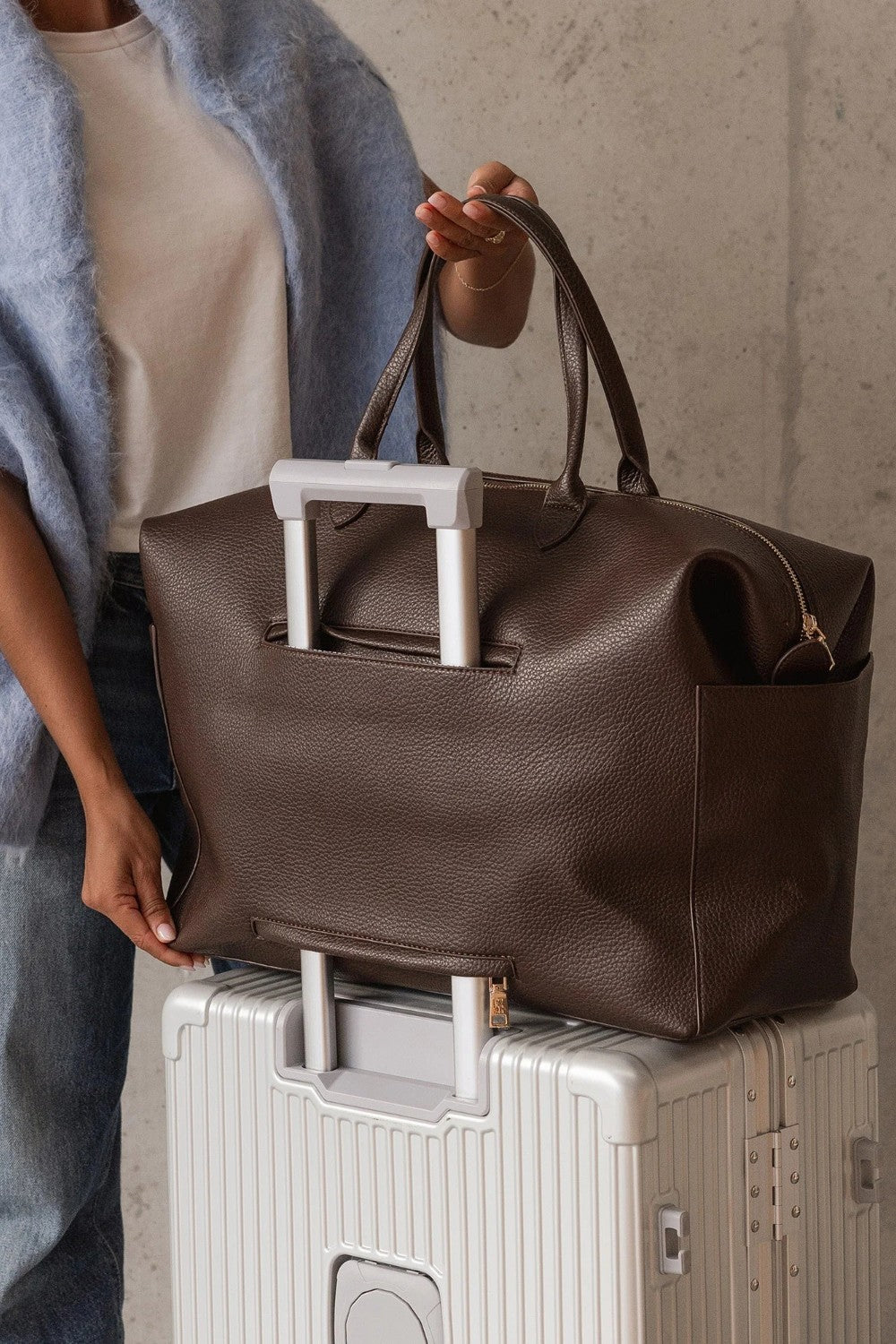A person wearing a light blue sweater and white top holds a large brown pebbled leather duffel bag with white handles, resting it on top of a silver rolling suitcase against a textured concrete wall.