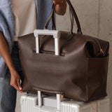 A person wearing a light blue sweater and white top holds a large brown pebbled leather duffel bag with white handles, resting it on top of a silver rolling suitcase against a textured concrete wall.