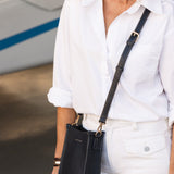 Person wearing a white shirt and white pants with a navy handbag, standing in front of a blurred background.