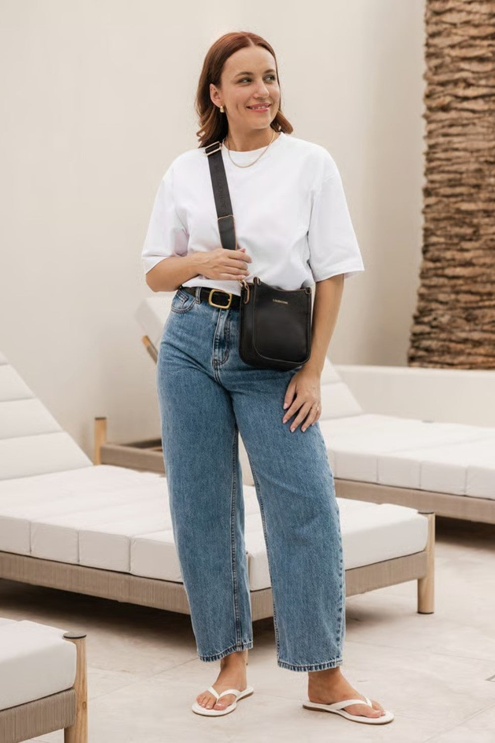 A woman wearing a white top and blue jeans carries a black leather crossbody bag with a matching strap in an indoor setting.
