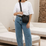 A woman wearing a white top and blue jeans carries a black leather crossbody bag with a matching strap in an indoor setting.