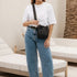 A woman wearing a white top and blue jeans carries a black leather crossbody bag with a matching strap in an indoor setting.
