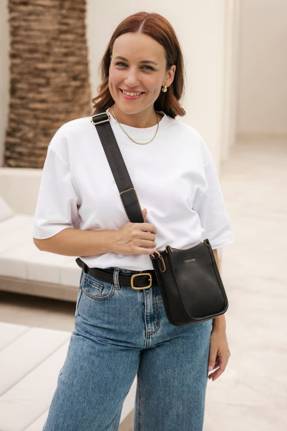 A woman wearing a white t-shirt and blue jeans models a black leather crossbody bag with a gold buckle and strap details.