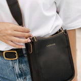 A woman wearing a white top and blue jeans carries a black leather crossbody bag with a matching strap.