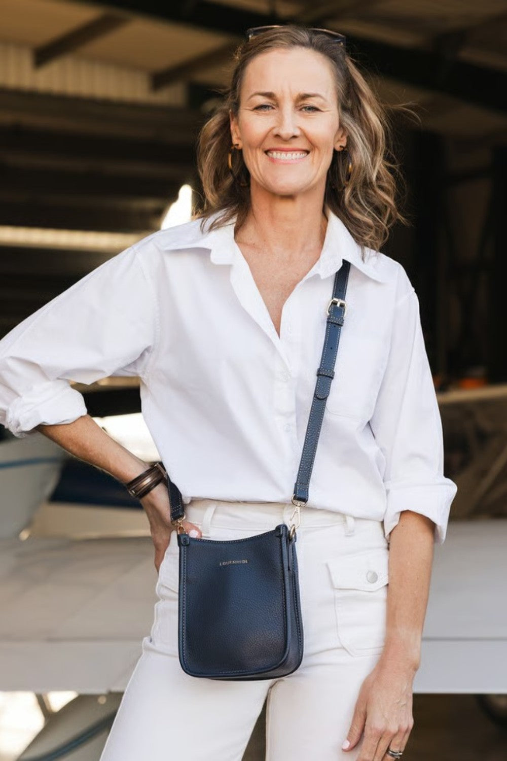 A woman wearing a white button-down shirt and white pants carries a navy blue leather crossbody bag with a matching strap in a boat garage setting.