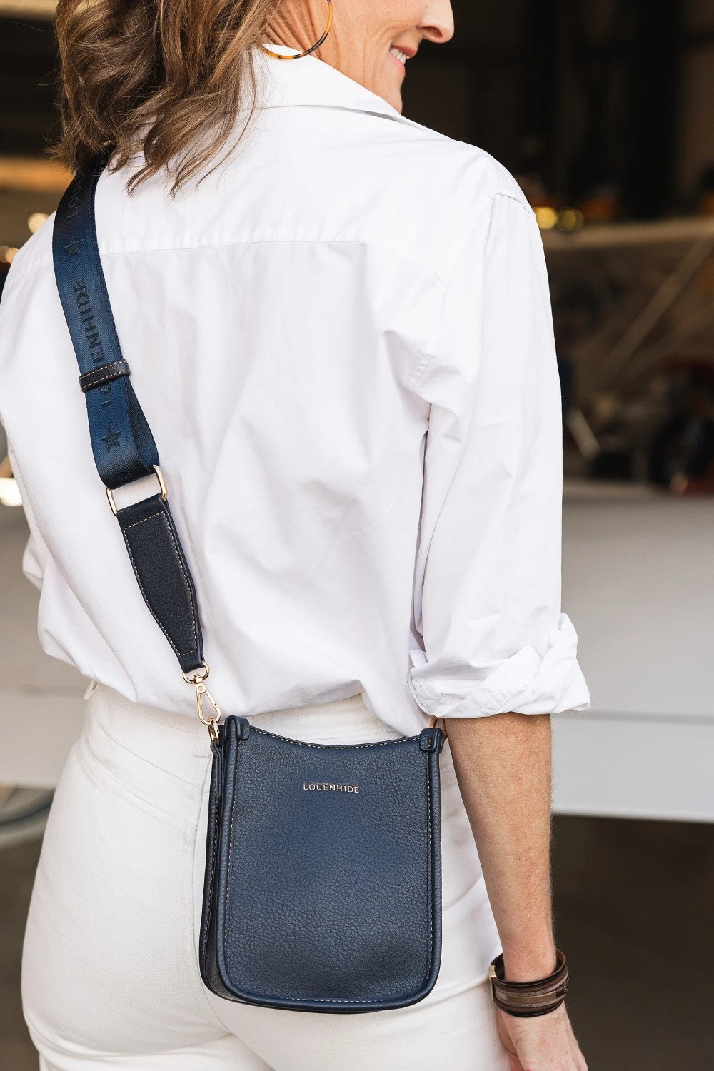 A woman wearing a white button-down shirt and white pants carries a navy blue textured leather crossbody bag with a matching navy strap. The bag has a small gold-tone logo reading 'LOUENHIDE'