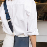 A woman wearing a white button-down shirt and white pants carries a navy blue textured leather crossbody bag with a matching navy strap. The bag has a small gold-tone logo reading 'LOUENHIDE'