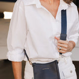A woman wearing a white button-down shirt and white pants carries a navy blue leather crossbody bag with a matching strap in a boat garage setting.