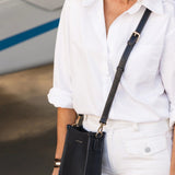A woman wearing a white button-down shirt and white pants carries a navy blue leather crossbody bag with a matching strap in a boat garage setting.