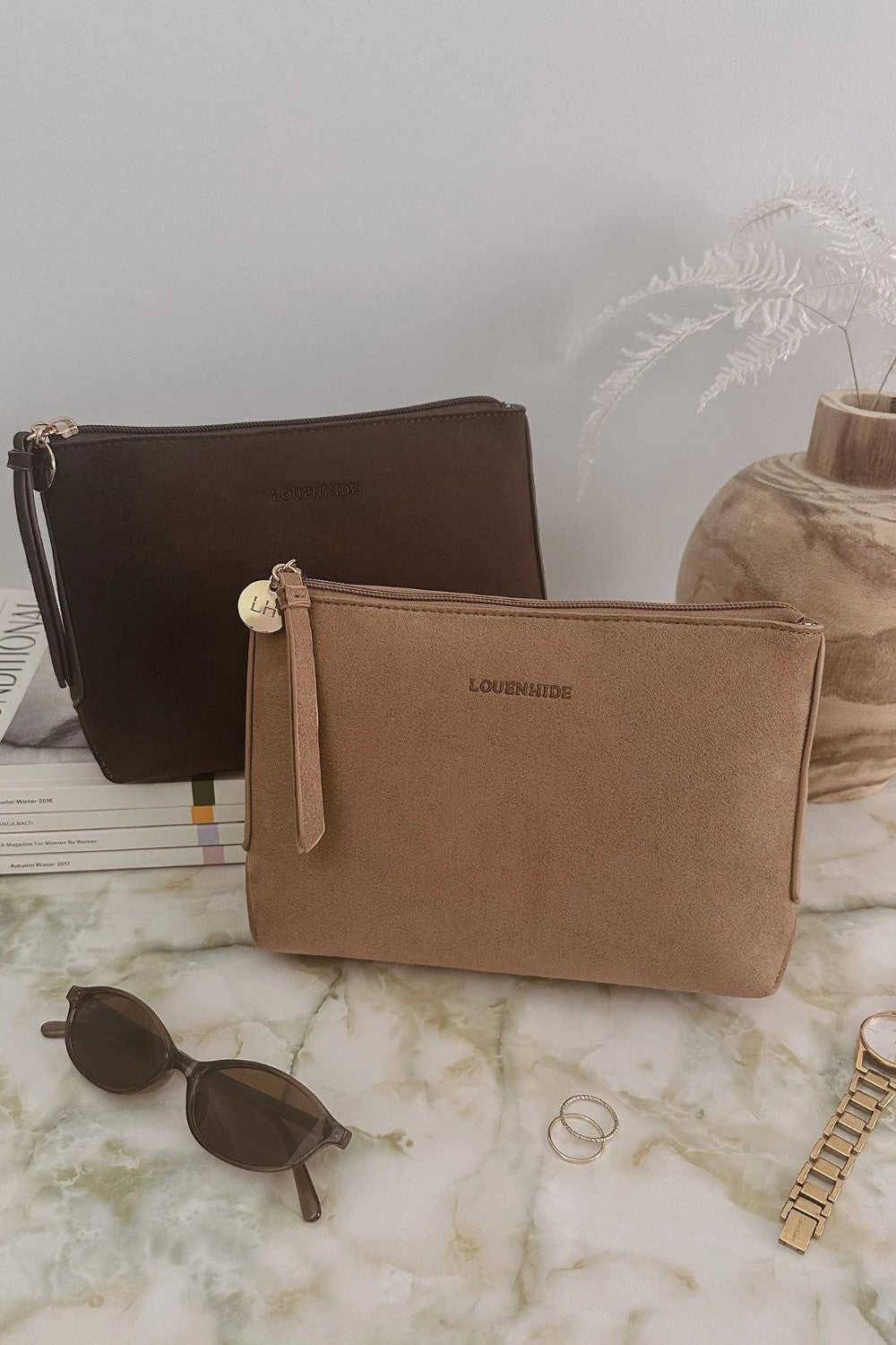 Two small zip pouches with wrist straps are displayed on a marble surface. The front pouch is tan and labeled LOUENHIDE, while the pouch behind it is dark brown.