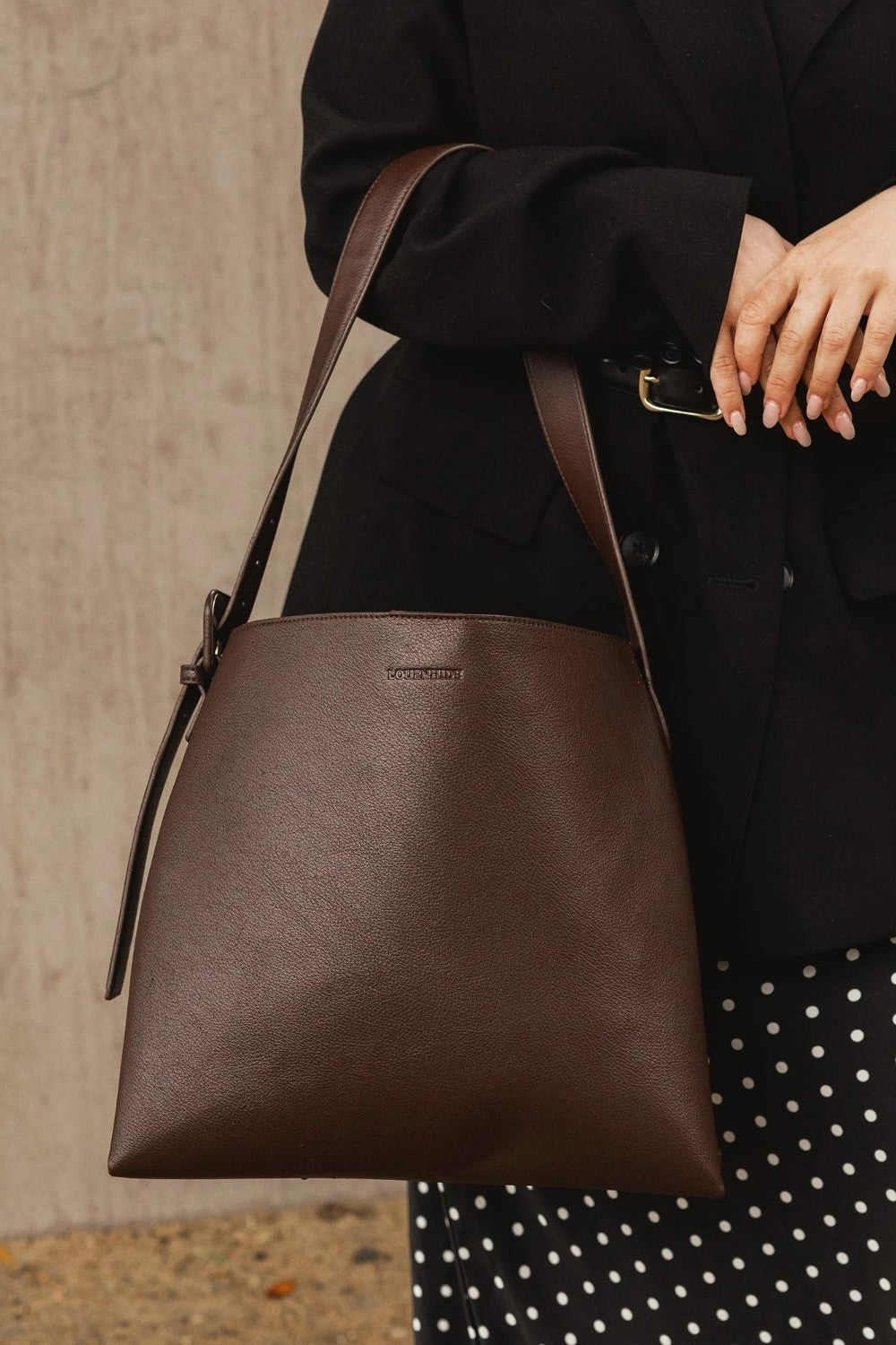 A person in a black blazer and black-and-white polka dot skirt carries a large brown leather shoulder bag with a matching strap against a textured beige wall.