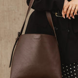 A person in a black blazer and black-and-white polka dot skirt carries a large brown leather shoulder bag with a matching strap against a textured beige wall.