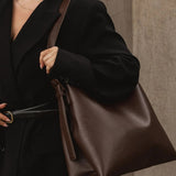 A woman in a black blazer carries a large brown leather shoulder bag with a wide strap, set against a neutral stone wall background.