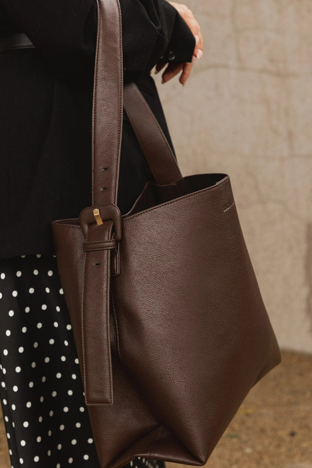A dark brown pebbled leather shoulder tote bag with a wide strap and a decorative buckle detail, shown being carried by a person wearing a black top and a black-and-white polka dot skirt.