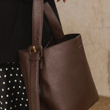 A dark brown pebbled leather shoulder tote bag with a wide strap and a decorative buckle detail, shown being carried by a person wearing a black top and a black-and-white polka dot skirt.