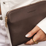 Brown leather clutch bag with gold zipper held by a person wearing a white jacket.
