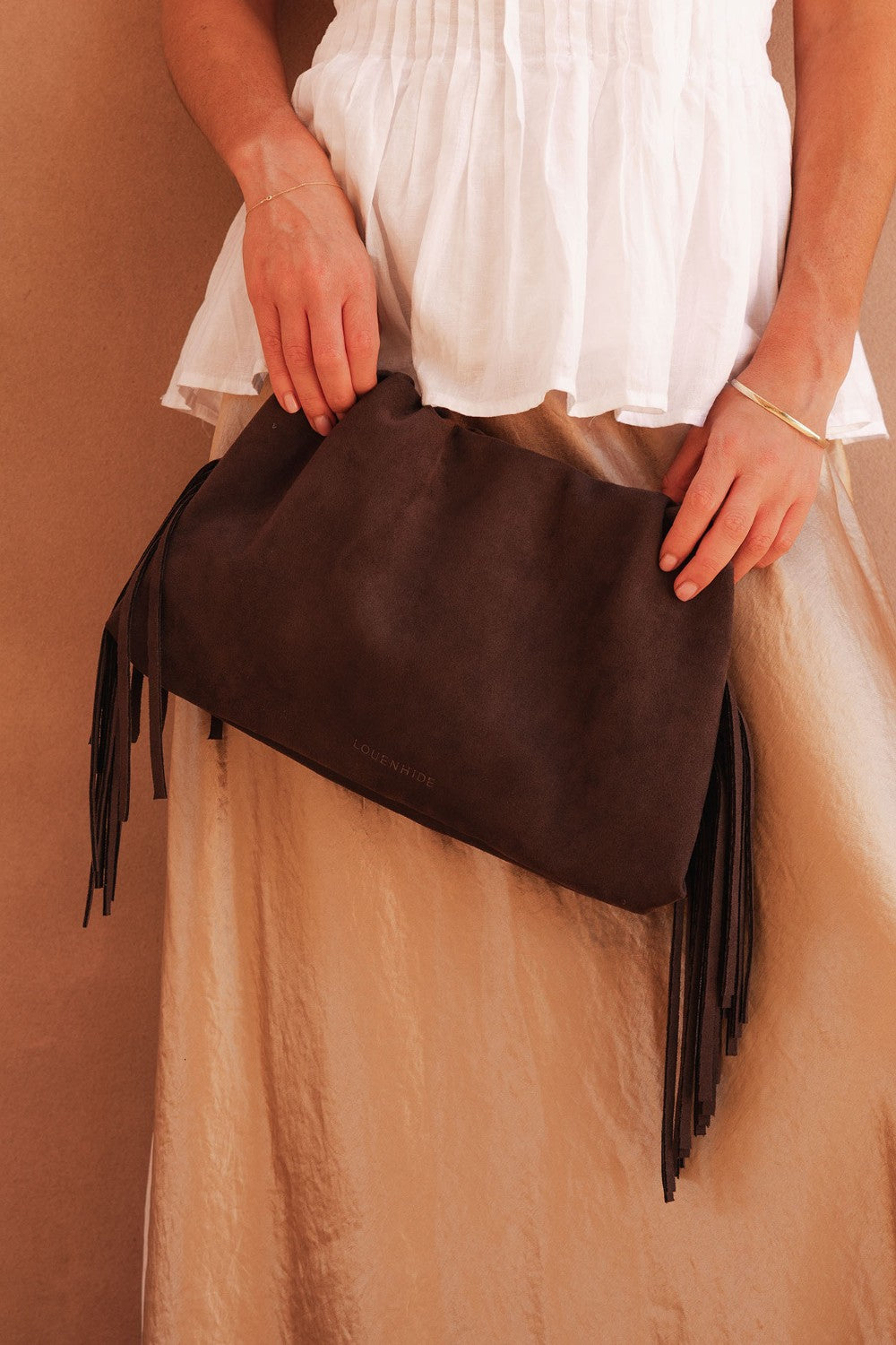 Brown clutch bag with fringes held by a person wearing a white top and beige skirt.