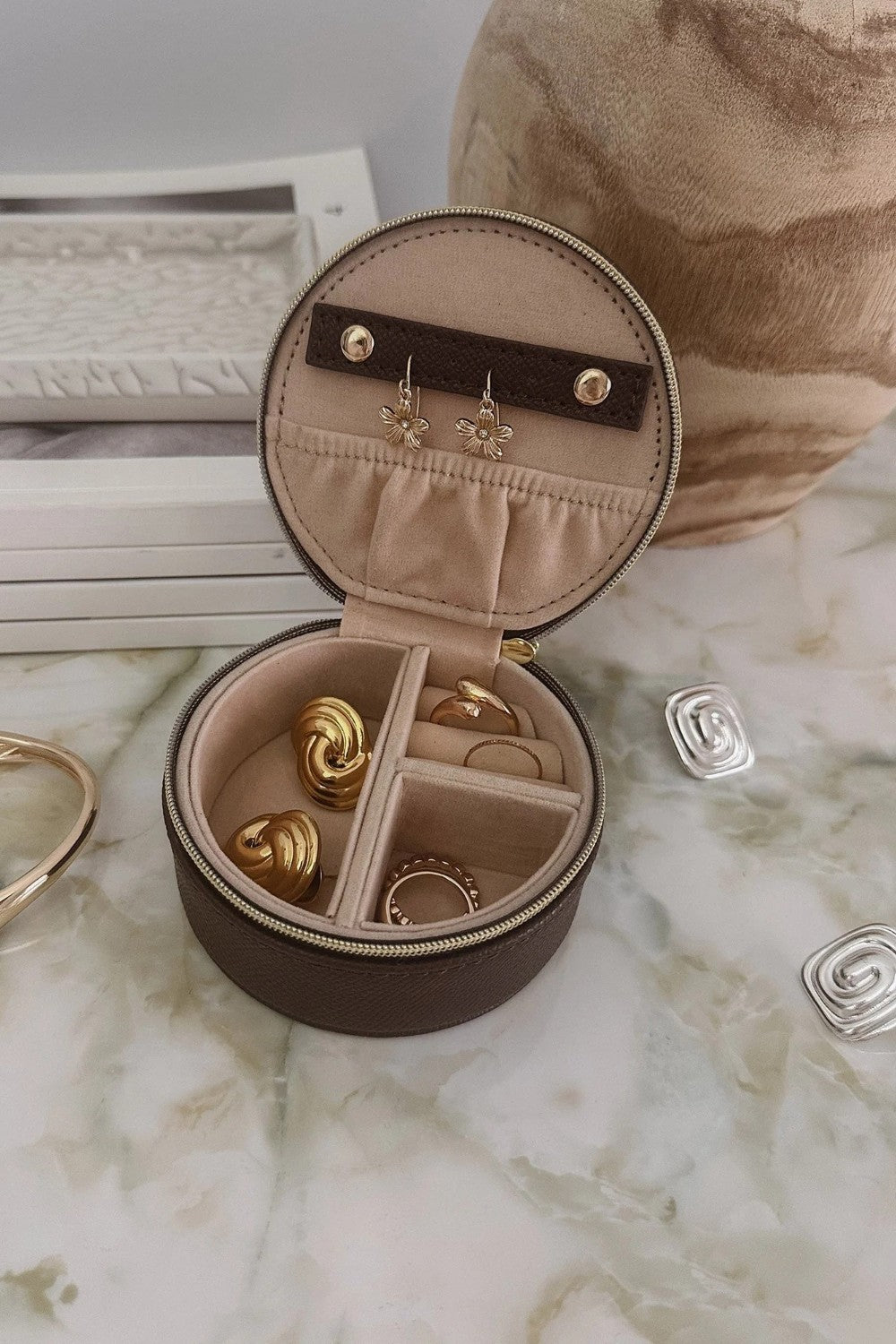 An open round jewelry organizer case with a dark brown exterior and beige interior, holding several gold-toned rings and earrings, styled on a marble-like surface with decorative props.