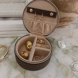 An open round jewelry organizer case with a dark brown exterior and beige interior, holding several gold-toned rings and earrings, styled on a marble-like surface with decorative props.
