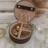 An open round jewelry organizer case with a dark brown exterior and beige interior, holding several gold-toned rings and earrings, styled on a marble-like surface with decorative props.