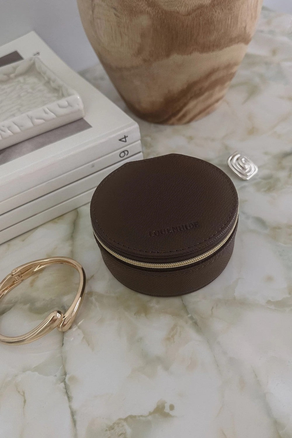 A small round dark brown leather jewelry box with a gold zipper, placed on a white marble surface next to gold and silver rings, with stacked books and a ceramic vase in the background.
