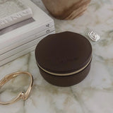 A small round dark brown leather jewelry box with a gold zipper, placed on a white marble surface next to gold and silver rings, with stacked books and a ceramic vase in the background.