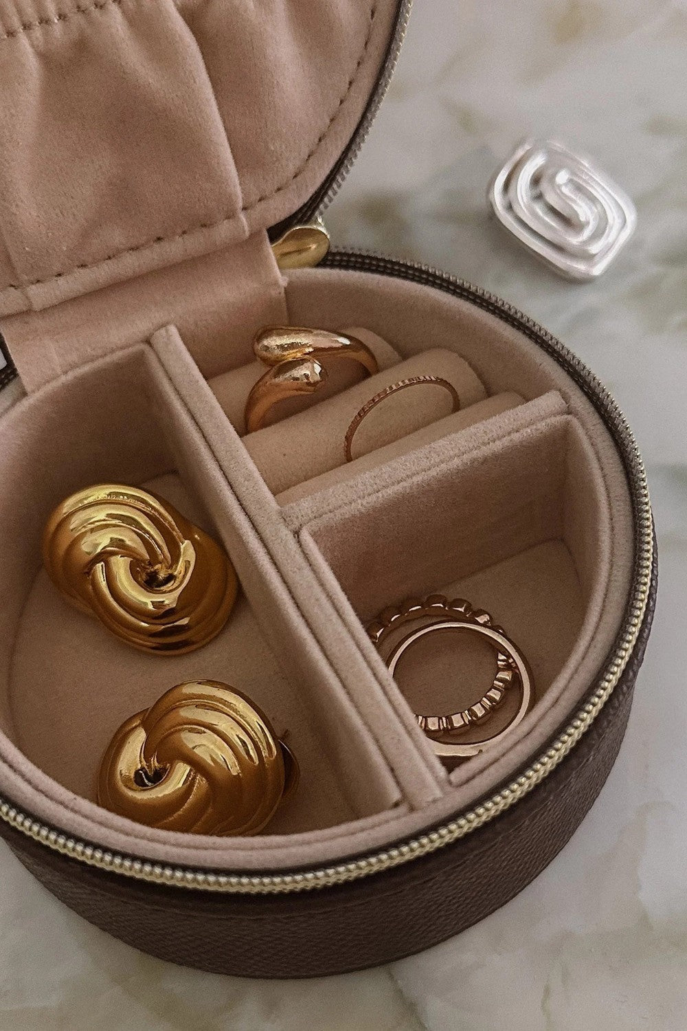 A round zippered jewelry organizer case with a beige velvet interior, shown open with several gold rings and earrings inside. A small silver ring is placed outside the case on a light marble-like surface.