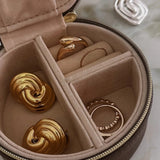 A round zippered jewelry organizer case with a beige velvet interior, shown open with several gold rings and earrings inside. A small silver ring is placed outside the case on a light marble-like surface.