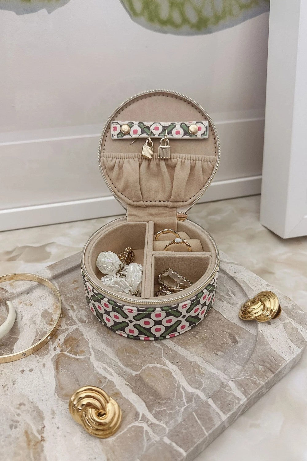 An open round jewelry box with a patterned exterior, beige interior compartments, styled on a marble tray with gold jewelry pieces.