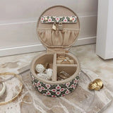 An open round jewelry box with a patterned exterior, beige interior compartments, styled on a marble tray with gold jewelry pieces.