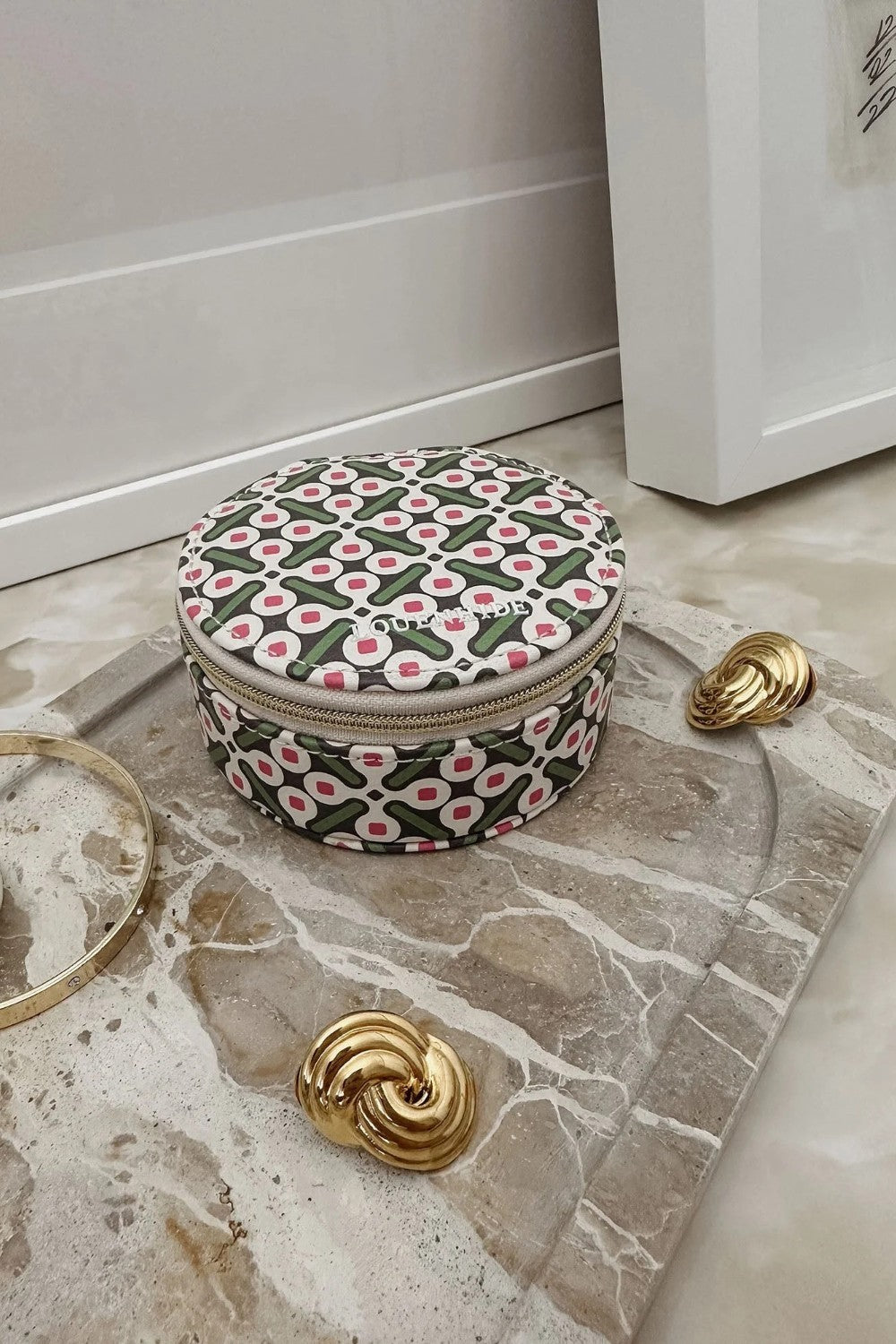 A round zippered jewelry box with a green, pink, and white geometric pattern, styled on a marble tray with gold accessories.