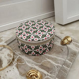 A round zippered jewelry box with a green, pink, and white geometric pattern, styled on a marble tray with gold accessories.