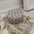 A round zippered jewelry box with a green, pink, and white geometric pattern, styled on a marble tray with gold accessories.
