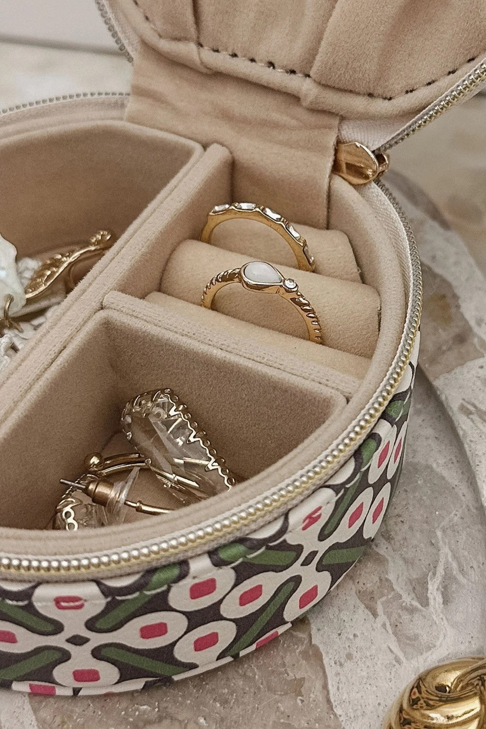 A round zippered jewelry organizer case with a green, pink, and white pattern, shown open to display its beige velvet interior compartments containing rings and earrings.