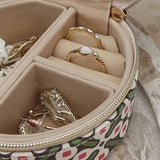 A round zippered jewelry organizer case with a green, pink, and white pattern, shown open to display its beige velvet interior compartments containing rings and earrings.